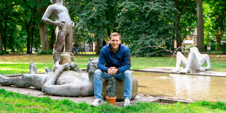 Auf dem Bild ist Jörn Flehinghaus zu sehen. Er sitzt auf einem Stein vor einem Brunnen. Vor ihm steht ein Kaktus, im Hintergrund sind eine Rasenfläche, Bäume und die Promenade zu sehen.<address>&copy; Uni MS – André Bednarz</address>
