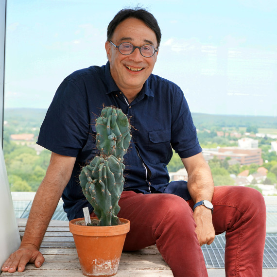 Das Bild zeigt Matthias Löwe. Neben ihm steht ein Kaktus, im Hintergrund sind von der Dachterrasse des UKM-Turms Gebäude und das Münsterland zu sehen.<address>&copy; Uni MS – Kathrin Nolte</address>