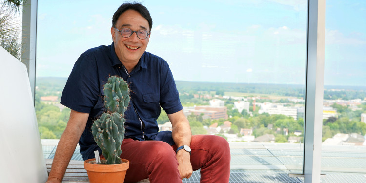 Das Bild zeigt Matthias Löwe. Neben ihm steht ein Kaktus, im Hintergrund sind von der Dachterrasse des UKM-Turms Gebäude und das Münsterland zu sehen.<address>&copy; Uni MS – Kathrin Nolte</address>