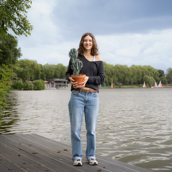 Das Bild zeit Julia Peris mit dem titelgebenden Kaktus in der Hand. Im Hintergrund ist der Aasee mit Segelbooten und Bäumen zu sehen.<address>&copy; Uni MS – André Bednarz</address>