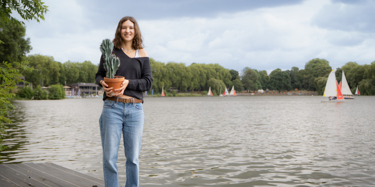 Das Bild zeit Julia Peris mit dem titelgebenden Kaktus in der Hand. Im Hintergrund ist der Aasee mit Segelbooten und Bäumen zu sehen.<address>&copy; Uni MS – André Bednarz</address>