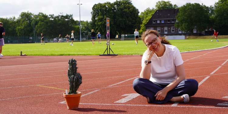 Barbara Halberschmidt in dunkler Jeans und weißen T-Shirt sitzt im Schneidersitz auf der Tartanbahn am Leonardo-Campus. Sie lächelt in die Kamera.<address>&copy; Uni MS - Kathrin Kottke</address>