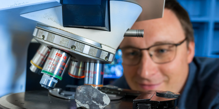 Das Bild zeigt im Vordergrund ein Mikroskop mit einem Meteoritenstück. Im Hintergrund ist Markus Patzek zu sehen, der das Fragment begutachtet.<address>&copy; Uni MS - Heiner Witte</address>