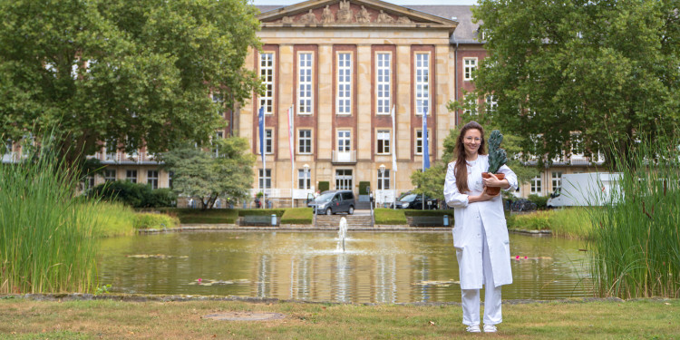 Das Foto zeigt Theresa Sonnenschein mit einem Kaktus in der Hand. Im Hintergrund sind Bäume, ein Teich und die Hautklinik zu sehen.<address>&copy; Uni MS - André Bednarz</address>
