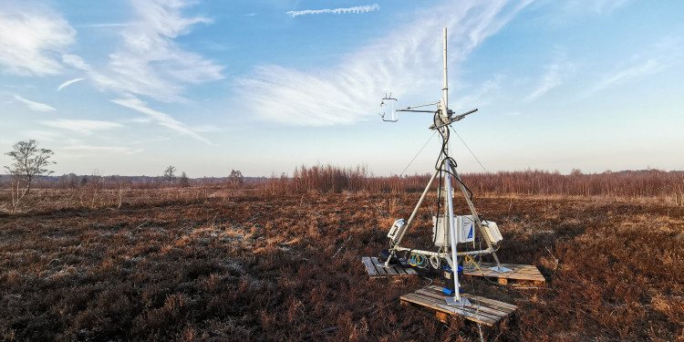 Auf einer weiten, flachen Moorlandschaft steht rechts im Bild ein silberfarbener Eddy-Kovarianz-Messturm. Er besteht aus einem etwa drei Meter hohen Metallgestell mit verschiedenen Messgeräten, Kabeln und Sensoren. Der Turm steht auf einer kleinen hölzernen Plattform. Im Hintergrund erstreckt sich eine offene, mit niedriger brauner Vegetation bewachsene Moorfläche. Am Horizont sind einzelne Bäume zu sehen, darüber ein weiter blauer Himmel mit dünnen weißen Wolken.<address>&copy; Biosphere-Atmosphere Interaction research group</address>
