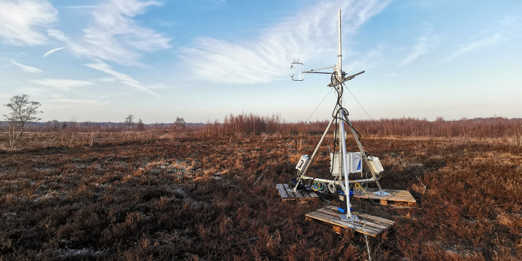 Set against a vast, flat moorland landscape, a silver-coloured Eddy covariance measurement tower stands on the right-hand side of the picture. It consists of a metal frame approximately three metres high, fitted with various measuring instruments, cables and sensors. The tower stands on a small wooden platform. In the background stretches an open moorland area covered with low brown vegetation. A few trees can be seen on the horizon, above which lies a vast blue sky dotted with thin white clouds.<address>&copy; Biosphere-Atmosphere Interaction research group</address>