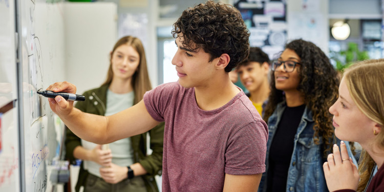 Ein Jugendlicher mit dunklen Locken und rotbraunem T-Shirt schreibt mit einem schwarzen Stift an ein Whiteboard. Um ihn herum stehen mehrere Mitschülerinnen und Mitschüler in einem hellen, modernen Klassenzimmer und verfolgen konzentriert, was er schreibt. Im Hintergrund sind Fenster und mit Aushängen bedeckte Wände zu erkennen.<address>&copy; stock.adobe.com - Rido</address>
