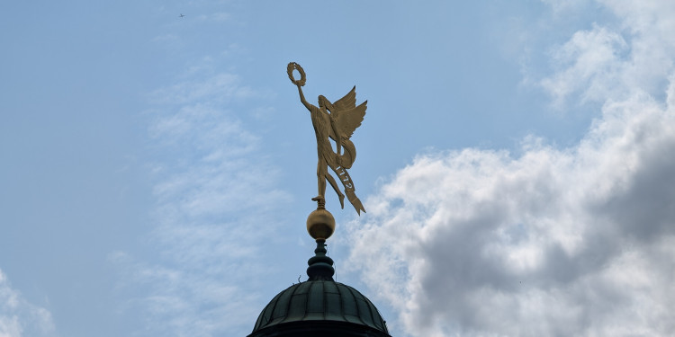 Auf dem Bild ist vor einem hellblauen, leicht bewölkten Himmel die bronzefarbene Nike-Figur auf der Kuppel des Münsteraner Schlosses zu sehen. Die geflügelte Figur steht auf einer Kugel und hält einen Lorbeerkranz empor.<address>&copy; Uni MS - Johannes Sträßer</address>