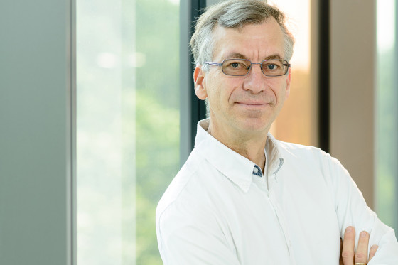Brustporträt: Ein Mann mit Brille und weißem Hemd steht mit verschränkten Armen in einem Raum. Im Hintergrund sieht man unscharf Fenster und dahinter das Grün von Bäumen.<address>&copy; Uni MS - Peter Dziemba</address>