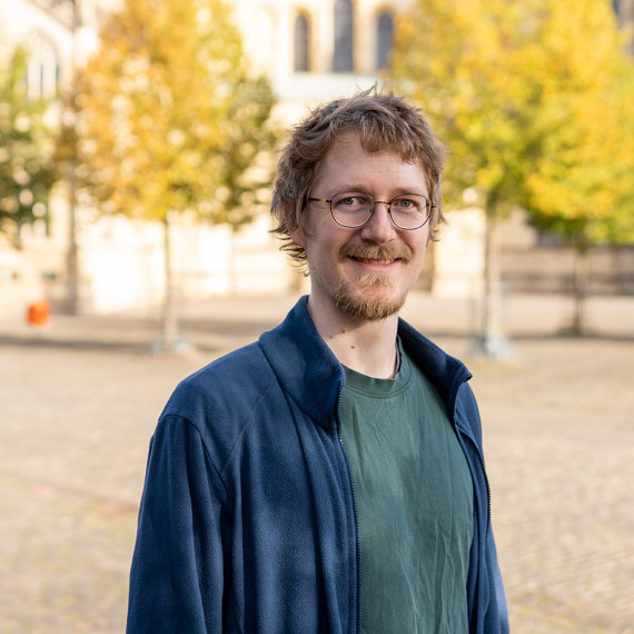 Ein Mann (Brustporträt) steht im Freien auf einem weiten, gepflasterten Platz. Der Hintergrund ist unscharf; man erkennt einige Bäume und den Teil einer Kirche/eines Doms.<address>&copy; Uni MS - Johannes Wulf</address>