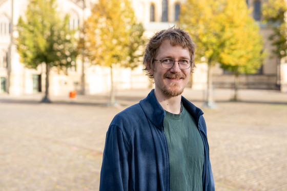 Ein Mann (Brustporträt) steht im Freien auf einem weiten, gepflasterten Platz. Der Hintergrund ist unscharf; man erkennt einige Bäume und den Teil einer Kirche/eines Doms.<address>&copy; Uni MS - Johannes Wulf</address>