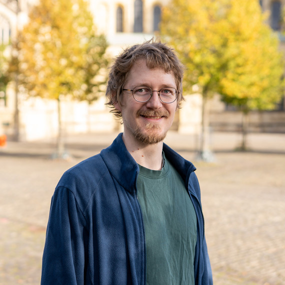 A man (chest-length portrait) is standing outdoors in a large, paved square. The background is out of focus; one can make out a few trees and part of a church or cathedral.<address>&copy; Uni MS - Johannes Wulf</address>
