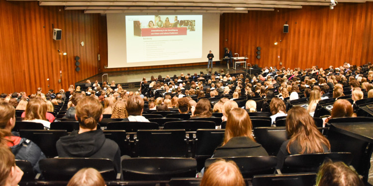 Großer Hörsaal mit zahlreichen Studierenden, die einer Präsentation auf einer Leinwand folgen. Auf der Bühne stehen zwei Personen.<address>&copy; Uni MS - Linus Peikenkamp</address>
