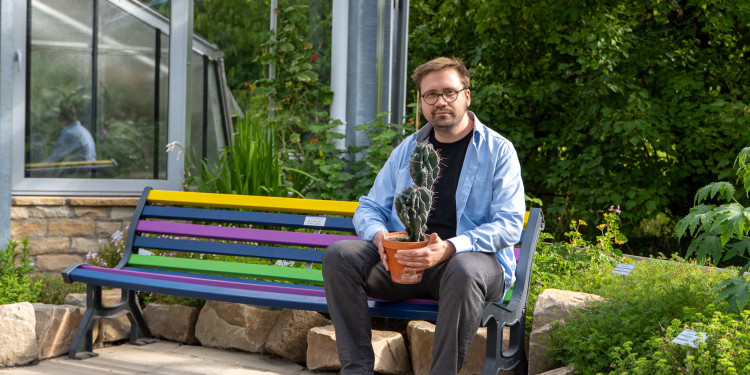 Das Bild zeigt Julius Jeiter auf einer bunten Bank sitzend. In seinen Händen hält er einen Topf mit einem Kaktus. Im Hintergrund sind ein Gewächshaus und grüne Pflanzen zu sehen.<address>&copy; Uni MS - André Bednarz</address>