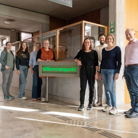 Das Bild, aufgenommen im Eingangsbereich des „Botanicums“, zeigt in der Mitte einen Leuchtschriftzug mit dem Wort „Willkommenspunkt“. Jeweils vier Personen stehen links und rechts vom Schriftzug und schauen in die Kamera.<address>&copy; Uni MS - André Bednarz</address>