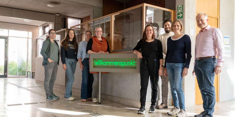 Das Bild, aufgenommen im Eingangsbereich des „Botanicums“, zeigt in der Mitte einen Leuchtschriftzug mit dem Wort „Willkommenspunkt“. Jeweils vier Personen stehen links und rechts vom Schriftzug und schauen in die Kamera.<address>&copy; Uni MS - André Bednarz</address>