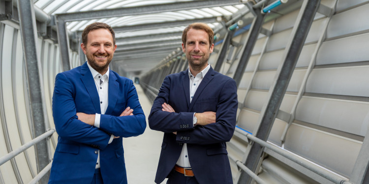 Das Bild zeigt Dr. Tim Hellwig (links) und Dr. Maximilian Brinkmann im Technologiepark. Sie tragen dunkle Anzüge, stehen nebeneinander mit verschränkten Armen und schauen freundlich in die Kamera.<address>&copy; Uni MS - Linus Peikenkamp</address>