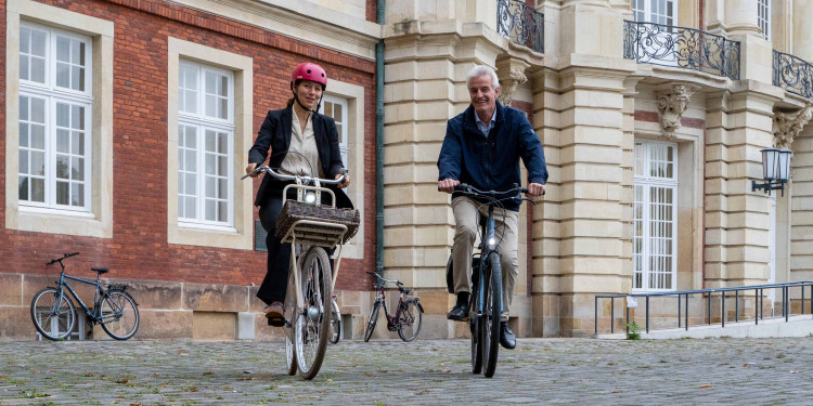 Das Bild zeigt Rektorin Prof. Dr. Susanne Menzel-Riedl und Pressesprecher Norbert Robers, die gemeinsam auf ihren Rädern vom Schloss wegfahren. Sie fahren direkt auf den Fotografen zu. Im Hintergrund ist die Hauptfassade des Schlosses zu sehen.<address>&copy; Uni MS - Johannes Wulf</address>