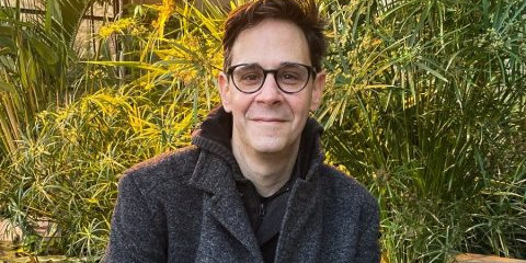 Ein Mann sitzt lächelnd auf einer niedrigen Mauer im Freien (im Hintergrund sind grüne Sträucher erkennbar).<address>&copy; privat</address>