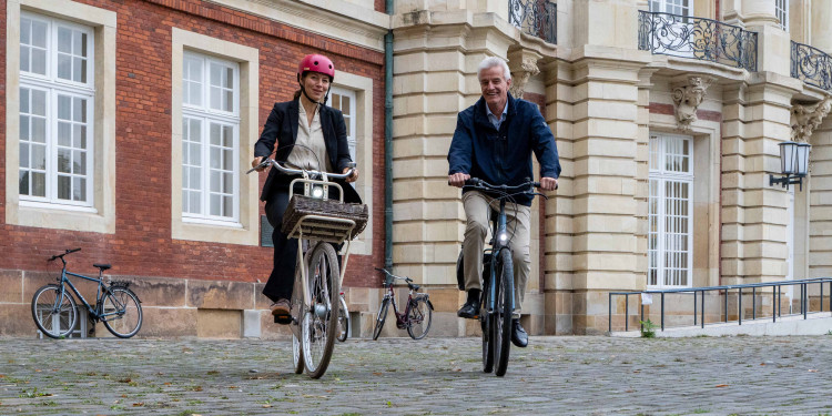 The image shows Rector Professor Dr Susanne Menzel-Riedl and press spokesperson Norbert Robers cycling away from the Schloss together. They are riding directly towards the photographer. The main facade of the Schloss can be seen in the background.<address>&copy; University of Münster - Johannes Wulf</address>