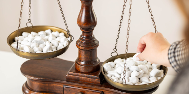 The image shows a balance scale with numerous small, white stones on each pan. A hand is adding another stone.<address>&copy; University of Münster - Roland Baege</address>