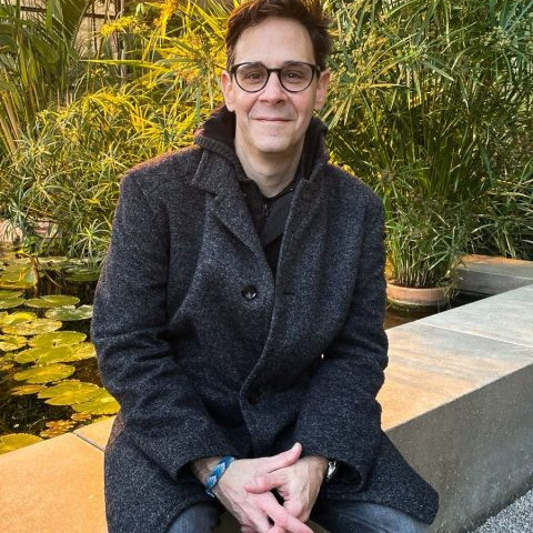 A man is sitting on a low wall outdoors, smiling (green bushes can be seen in the background).<address>&copy; private</address>