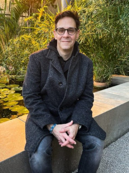 A man is sitting on a low wall outdoors, smiling (green bushes can be seen in the background).<address>&copy; private</address>