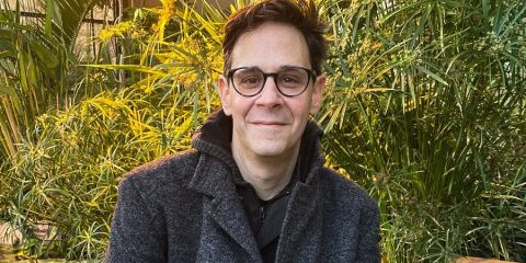 A man is sitting on a low wall outdoors, smiling (green bushes can be seen in the background).<address>&copy; private</address>
